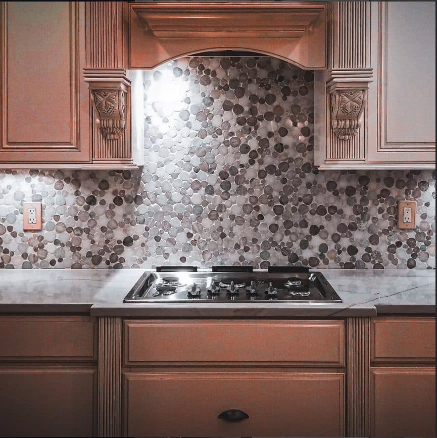 Stone mosaic backsplash behind a cooktop with wood range hood and cabinets